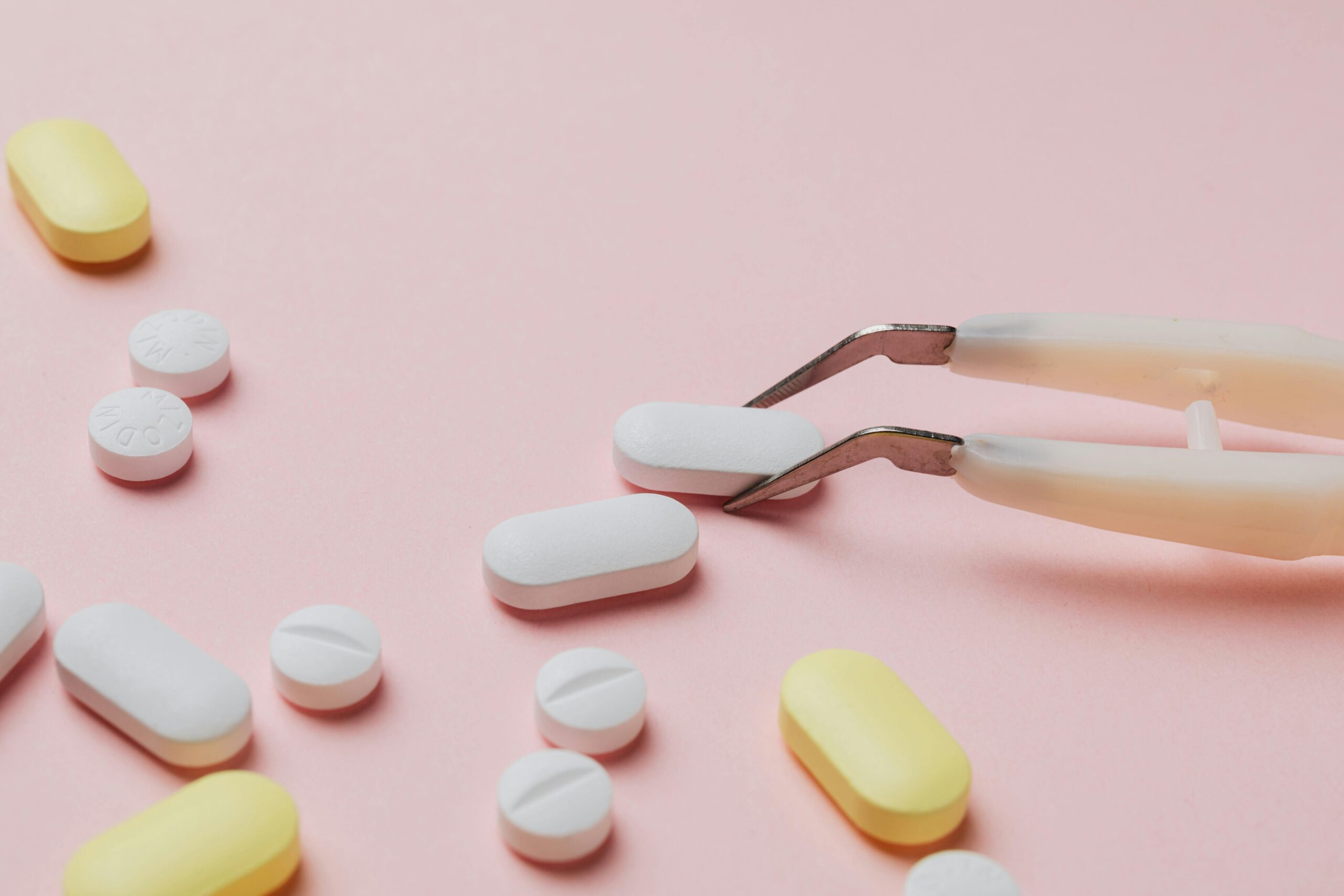 Different pills and tablets on pastel pink background with tweezers.